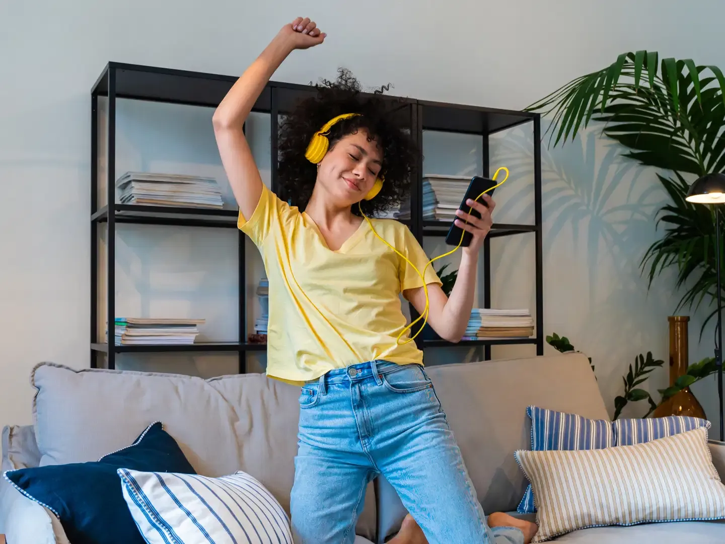 A cheerful woman wearing yellow headphones kneeling and dancing on a sofa while holding a smartphone.